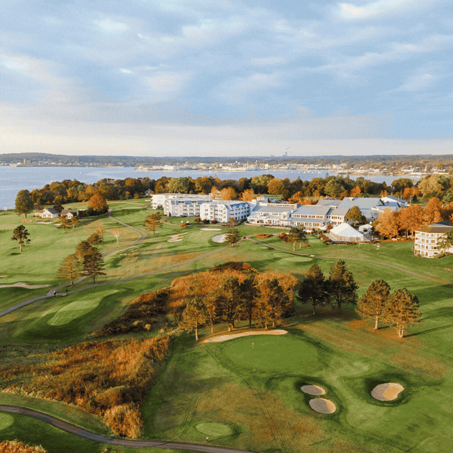 Aerial view of a golf course with well-manicured greens, sand bunkers, and trees in autumn colors. Buildings and a body of water are visible in the background under a partly cloudy sky.