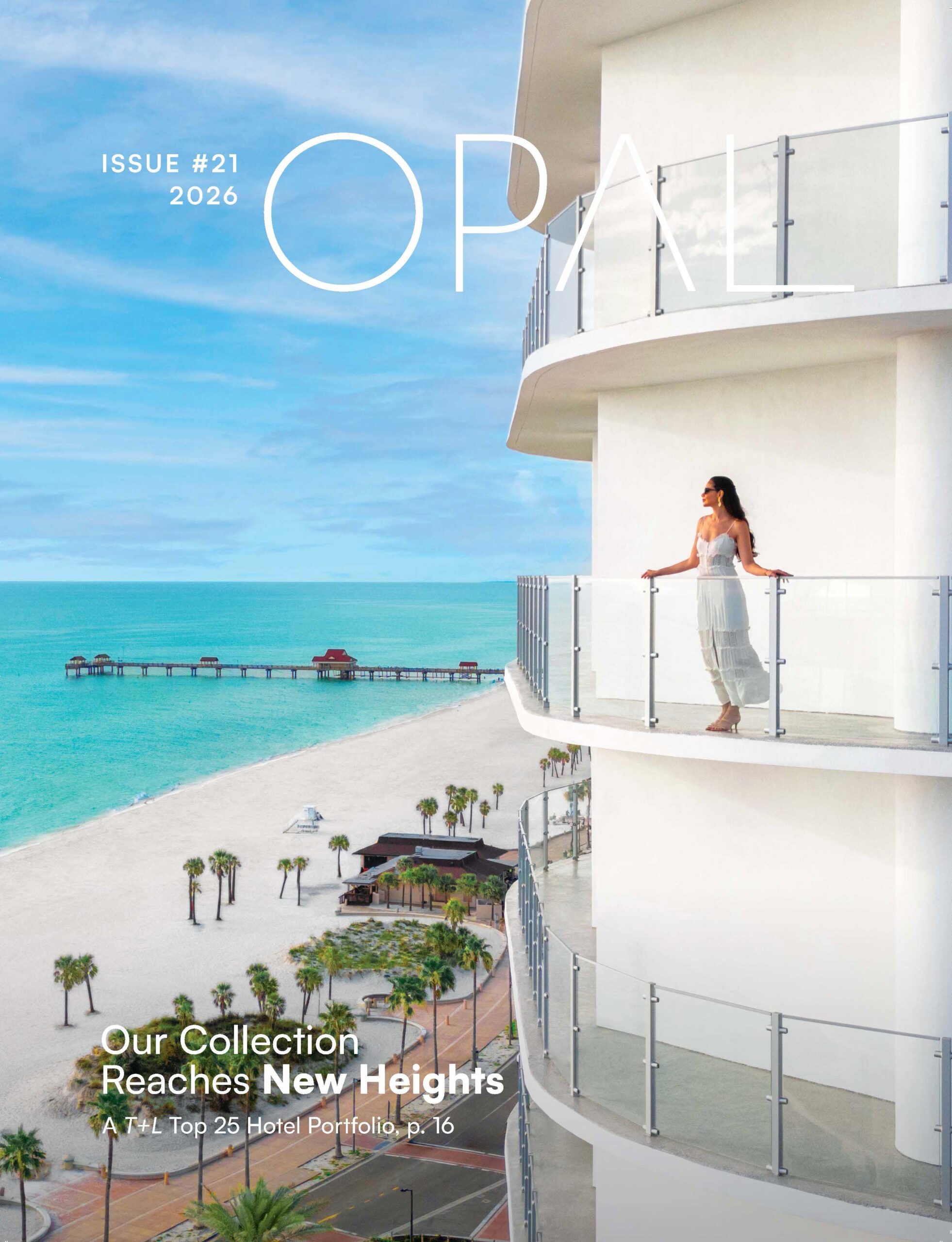 A woman in a white dress stands on a curved balcony of a modern hotel overlooking a sandy beach, palm trees, and turquoise ocean under a clear sky. The cover reads OPAL Issue #21 2026 and features headlines in white text.