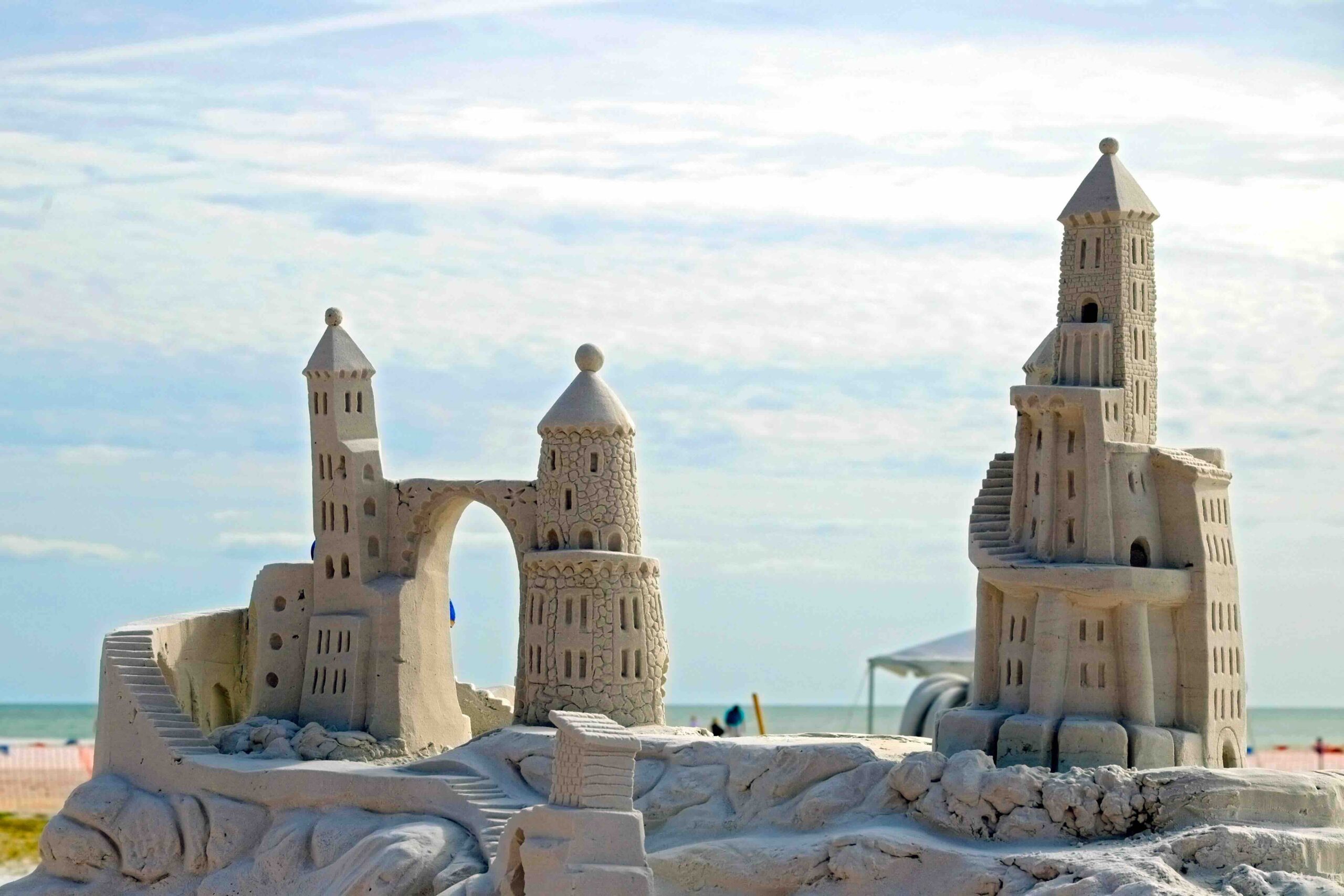A detailed sandcastle sculpture with towers, domes, and arches stands on a beach under a bright sky, with the ocean and distant people in the background.