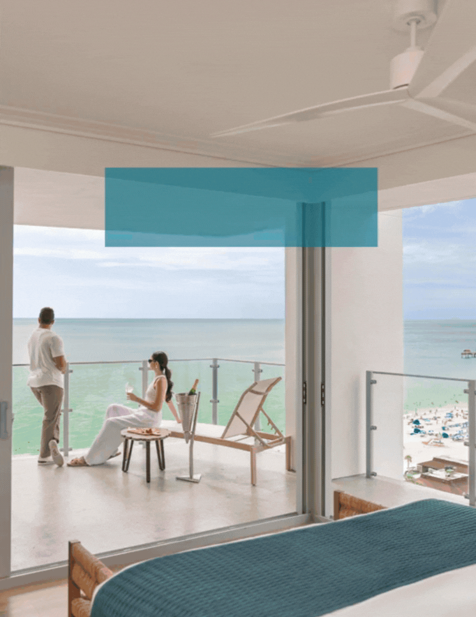 A man and woman relax on a balcony overlooking the ocean, with beachgoers visible below. The balcony has lounge chairs and a table with champagne, seen from inside a modern, bright bedroom.
