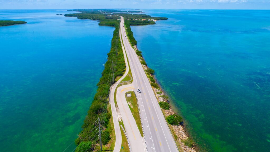 Aerial view of a long, narrow road surrounded by turquoise ocean waters, connecting small islands with green vegetation under a bright blue sky. A few vehicles travel along the road.