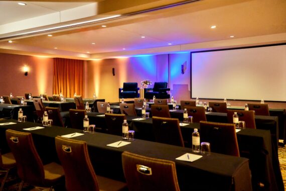 A conference room with rows of tables and chairs facing a stage with two armchairs, a small table, and a projector screen. Each table has notepads, pens, glasses, and bottled water. Warm lighting fills the room.