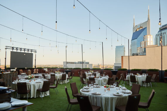 A rooftop event space with round tables set for dining, brown chairs, string lights overhead, and city skyscrapers, including the AT&T building, visible in the background under a clear sky.