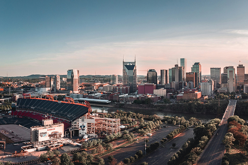 Aerial view of downtown Nashville, Tennessee, featuring the skyline with prominent skyscrapers, Nissan Stadium on the left, the Cumberland River, and tree-lined roads and parking areas in the foreground under a clear sky.