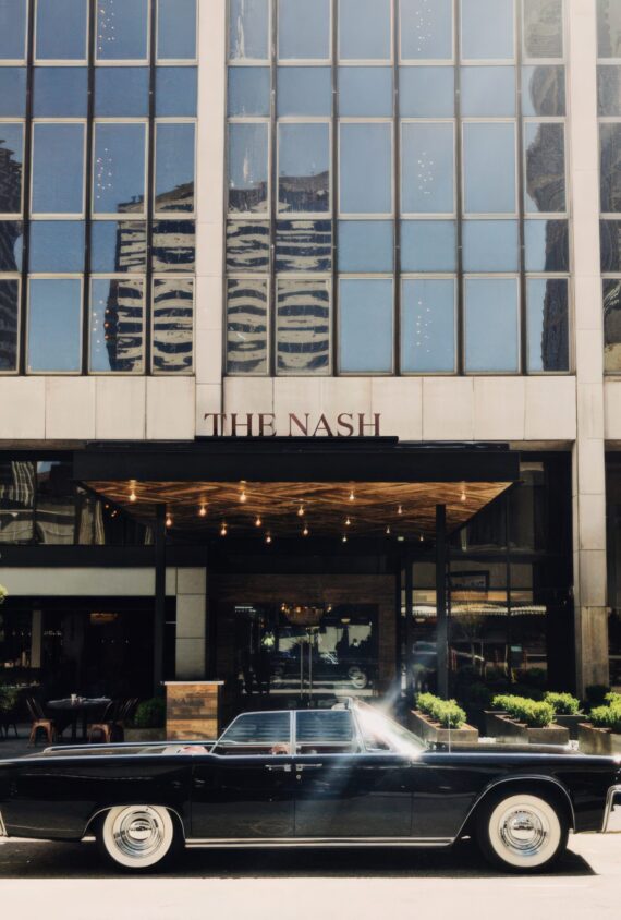 A vintage black car is parked in front of a modern glass building with a sign that reads THE NASH. The building reflects neighboring skyscrapers in its windows.