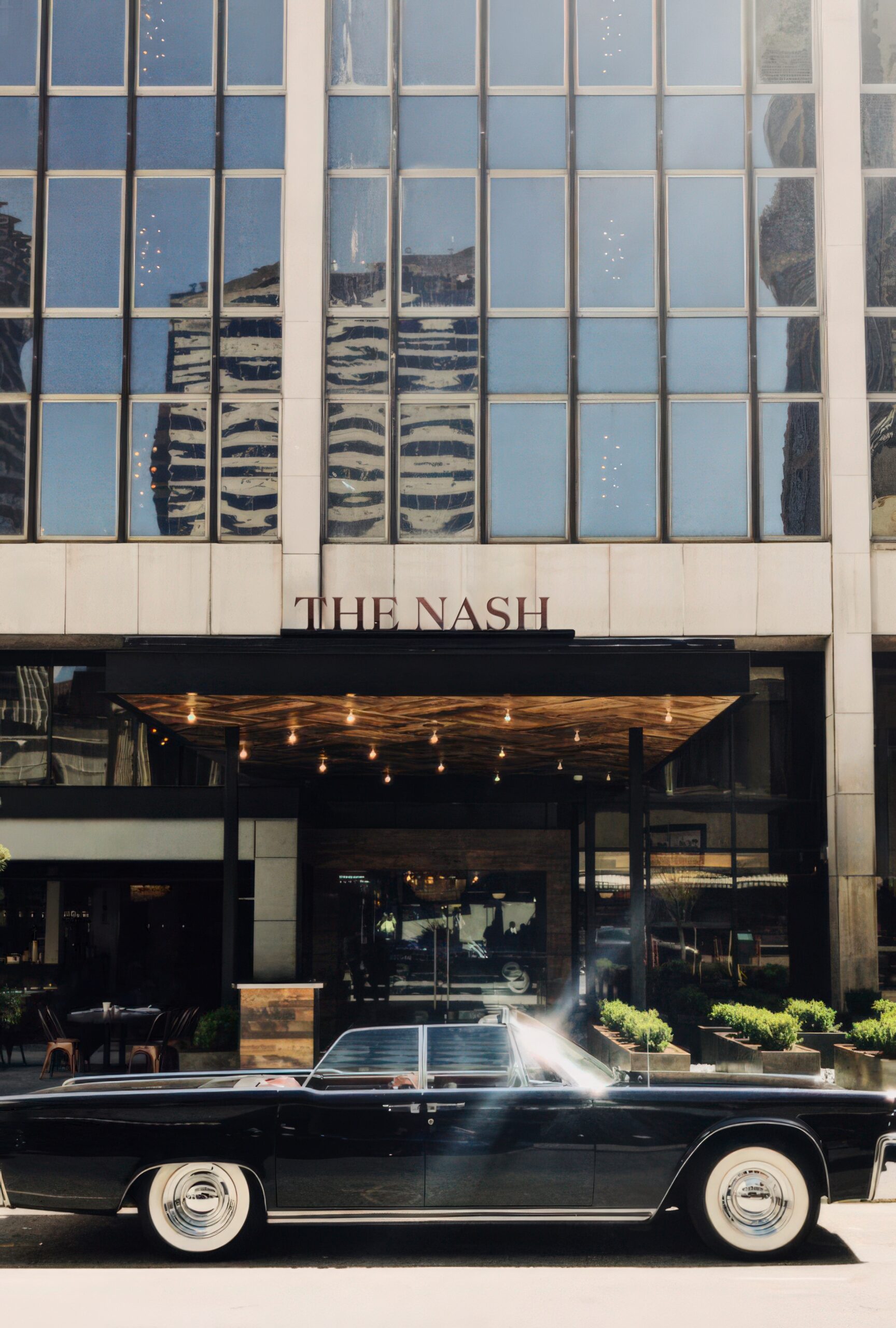 A vintage black car is parked in front of a modern glass building with a sign that reads THE NASH. The building reflects neighboring skyscrapers in its windows.