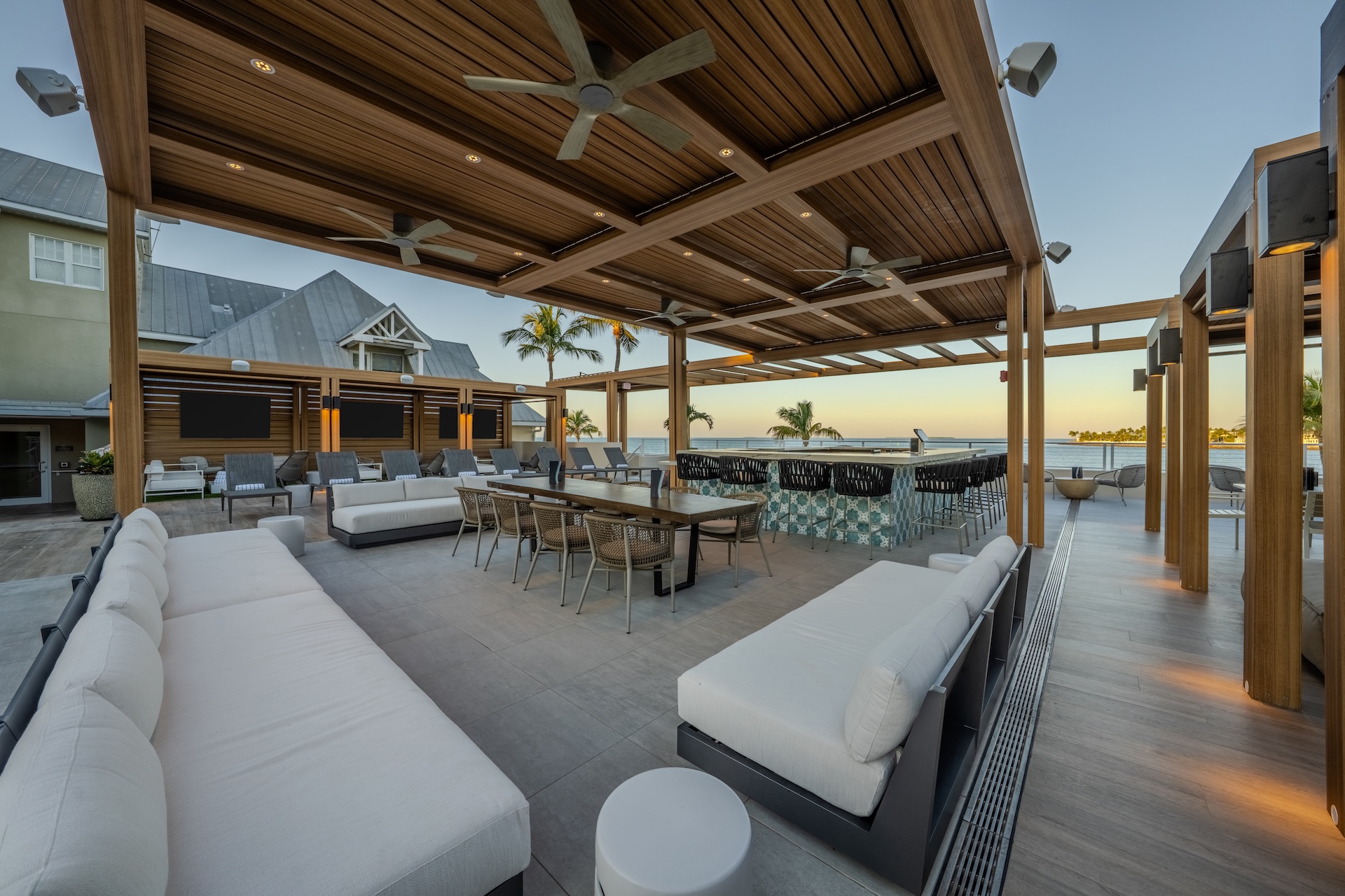 Modern outdoor lounge with wooden pergola, ceiling fans, white cushioned seating, dining tables, a bar with stools, and palm trees in the background—reminiscent of the most stylish rooftop bars—overlooking the ocean at sunset.