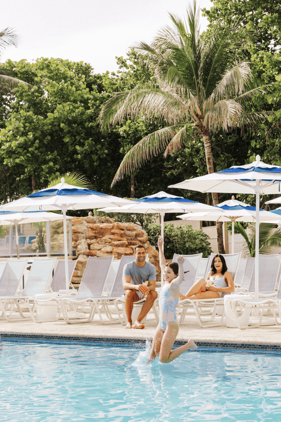 A young girl jumps into a swimming pool while two adults, seated on lounge chairs under blue and white umbrellas, watch and smile. Palm trees and lush greenery are in the background.