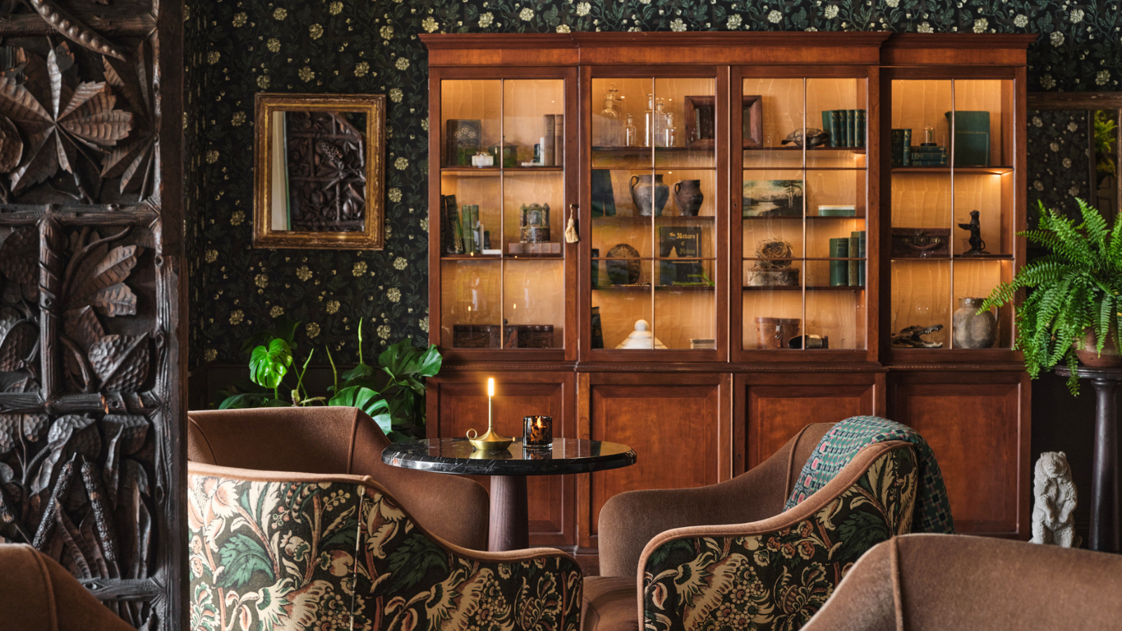 A cozy vintage room with floral wallpaper, patterned armchairs around a small round table with a lit candle, a large wooden display cabinet filled with books and artifacts, and green potted plants.