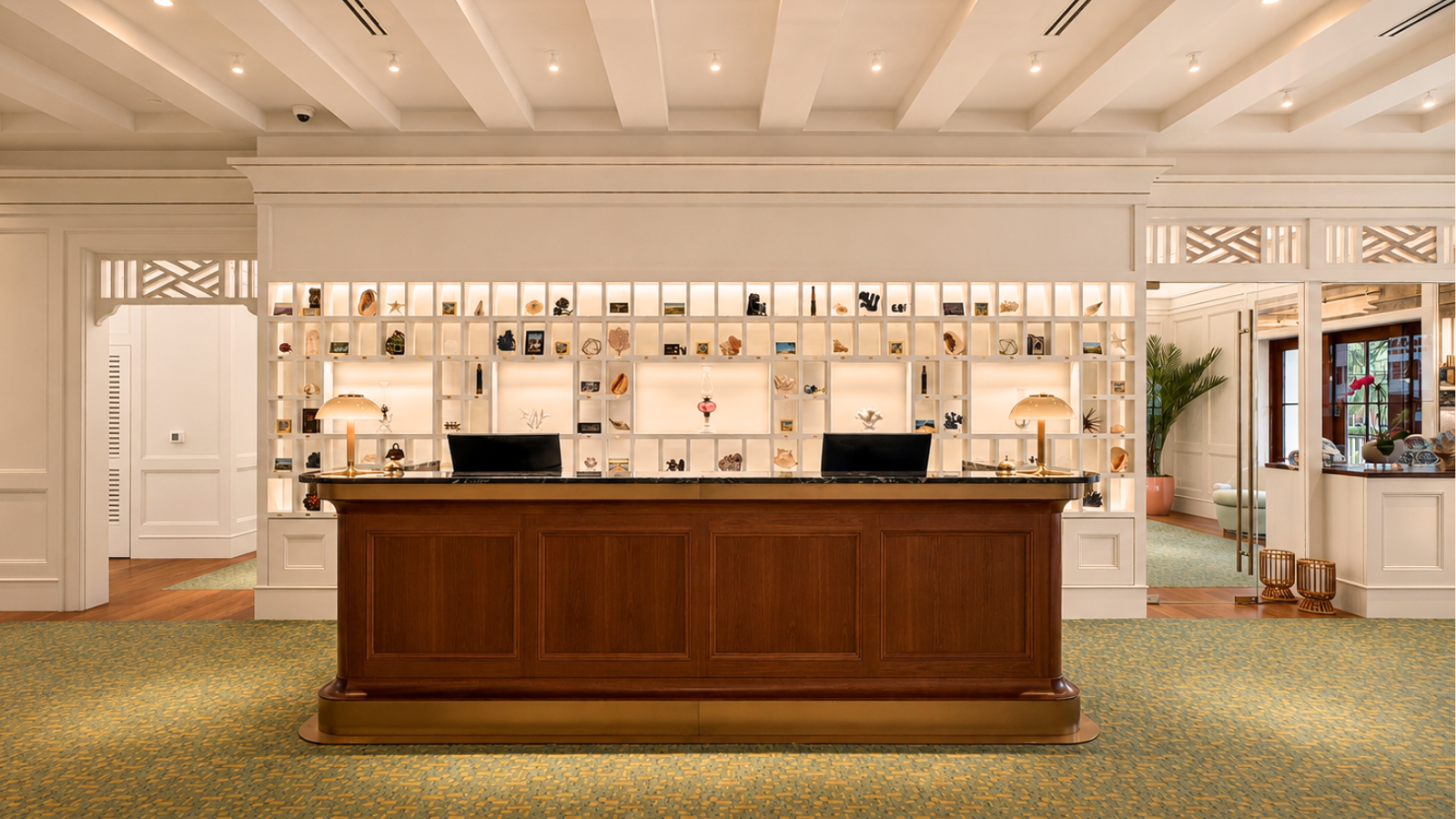 A hotel reception desk with a wooden counter, two computer monitors, and a decorative shelving wall behind displaying various ornaments and books. The area is well-lit with a patterned green carpet.