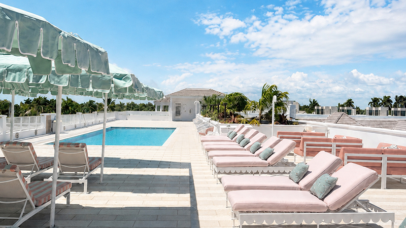 A rooftop pool area with light pink lounge chairs, green umbrellas, and palm trees under a blue sky with scattered clouds. White railings and a building are visible in the background.