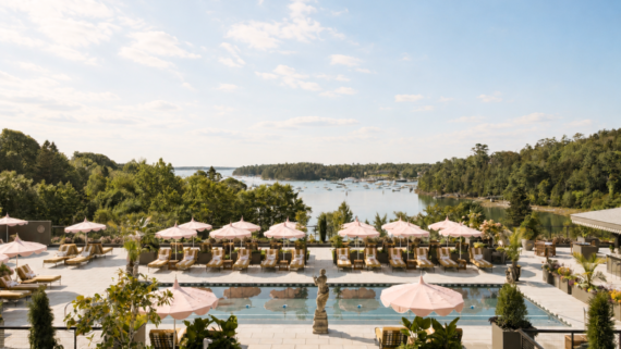A luxurious pool area with pink umbrellas and lounge chairs overlooks a peaceful harbor filled with boats, surrounded by lush greenery under a blue sky with scattered clouds.