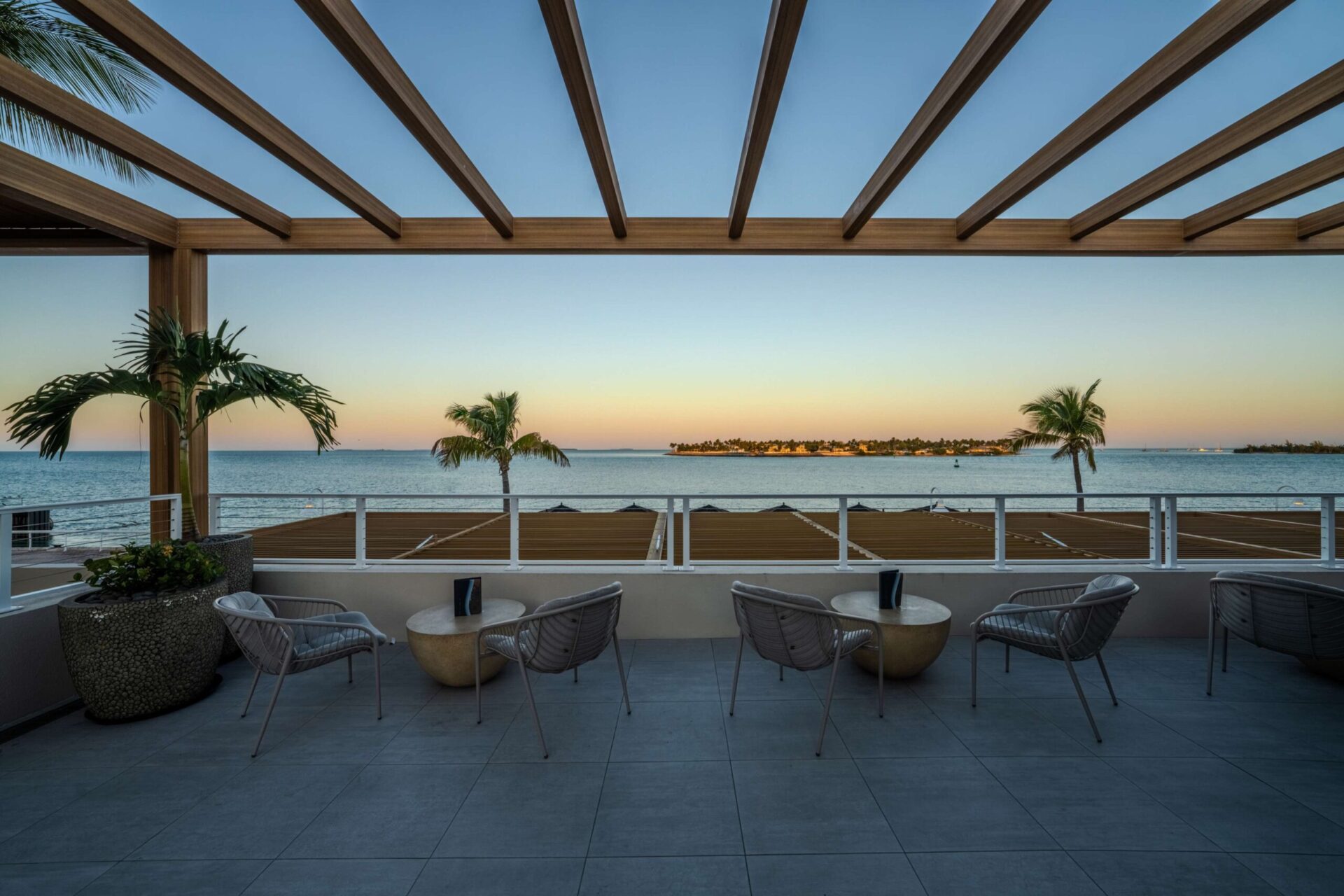 Outdoor patio with modern chairs and tables overlooking the ocean, palm trees, and a distant shoreline at sunset, framed by a wooden pergola overhead.