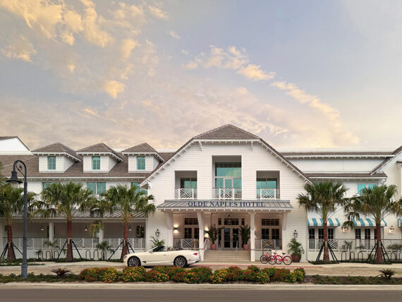 A white, coastal-style hotel with palm trees out front, a white convertible car parked by the entrance, and two red bicycles nearby, under a partly cloudy sky at sunset.