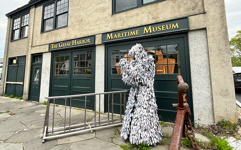 A statue covered in silvery feather-like shapes stands outside the Great Harbor Maritime Museum, next to a bike rack and weathered stone pavement—a striking landmark featured in the Northeast Harbor Guide: Best Activities & Attractions.