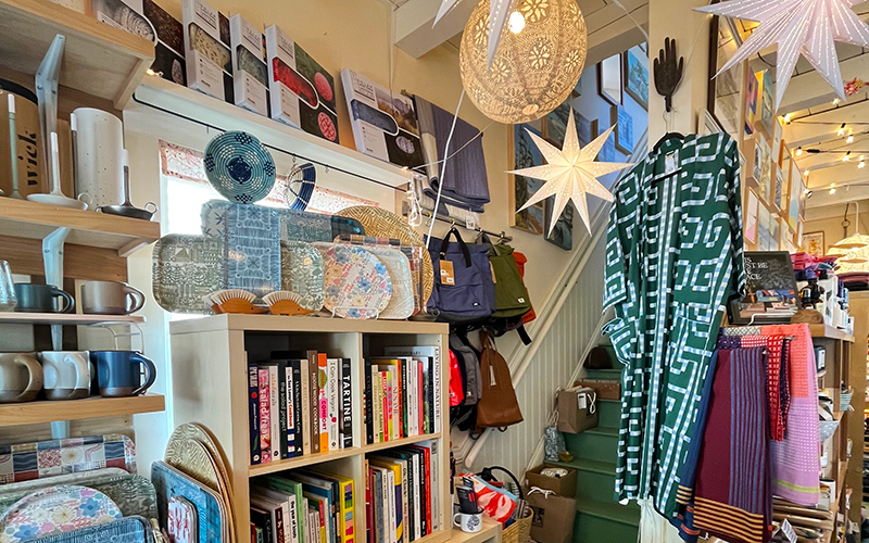 A cozy shop interior with books, mugs, trays, bags, and textiles neatly displayed invites exploration inspired by the Northeast Harbor Guide: Best Activities & Attractions. Hanging star-shaped lights and a patterned robe add charm, while shelves are filled with colorful and assorted household items reminiscent of local treasures found in Northeast Harbor.