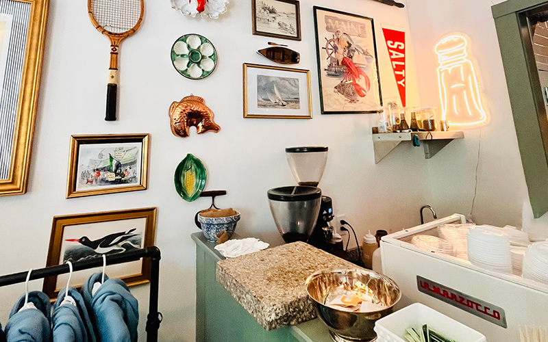 Cozy cafe corner with framed art, vintage tennis racket, green leaf plate, neon salt shaker sign, and coffee equipment on a marble counter next to stacked plates—evoking the charm you’ll find highlighted in the Northeast Harbor Guide: Best Activities & Attractions.