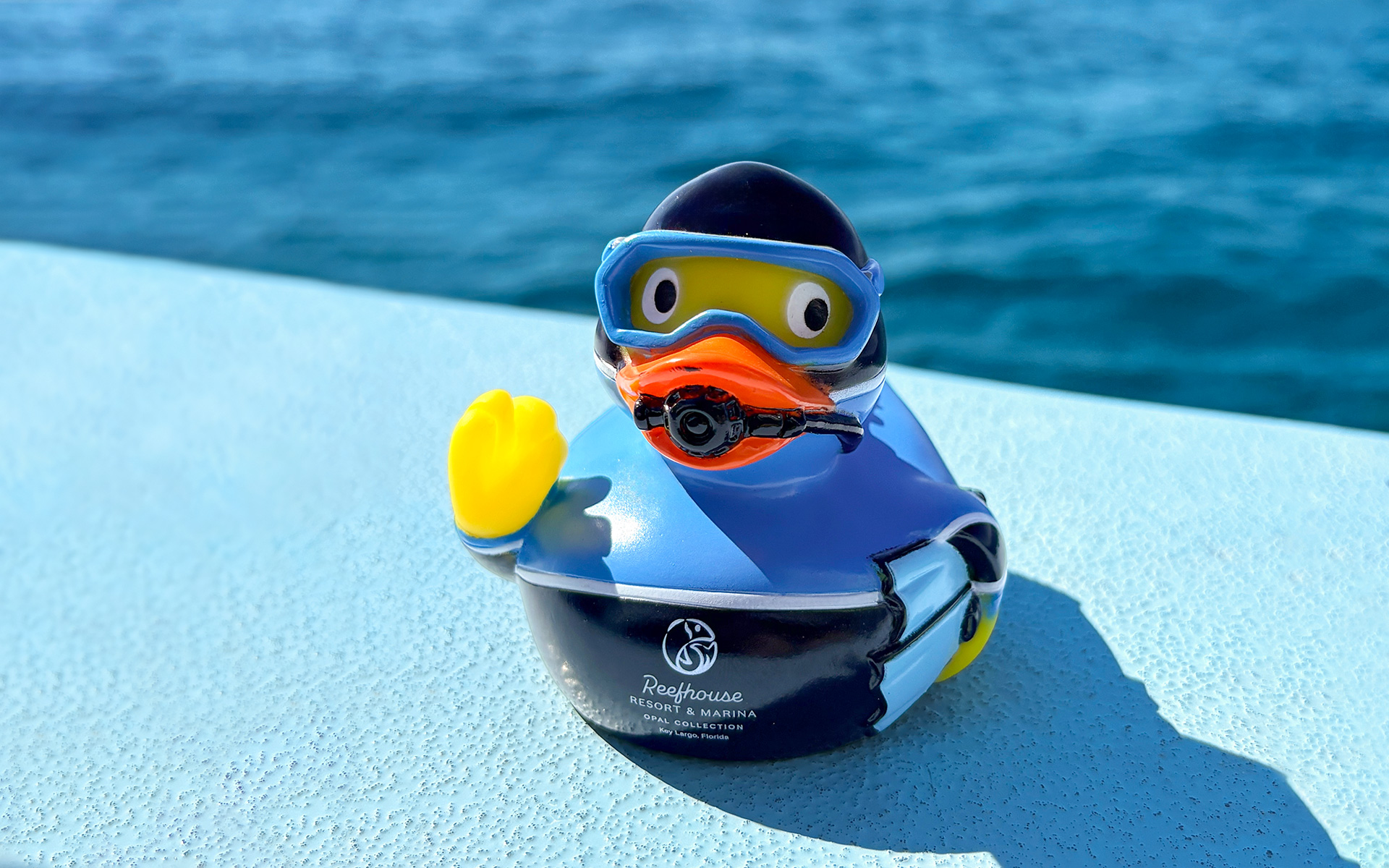 A rubber duck dressed as a scuba diver with the Reefhouse Resort & Marina logo, sitting on a boat deck with the ocean in the background.