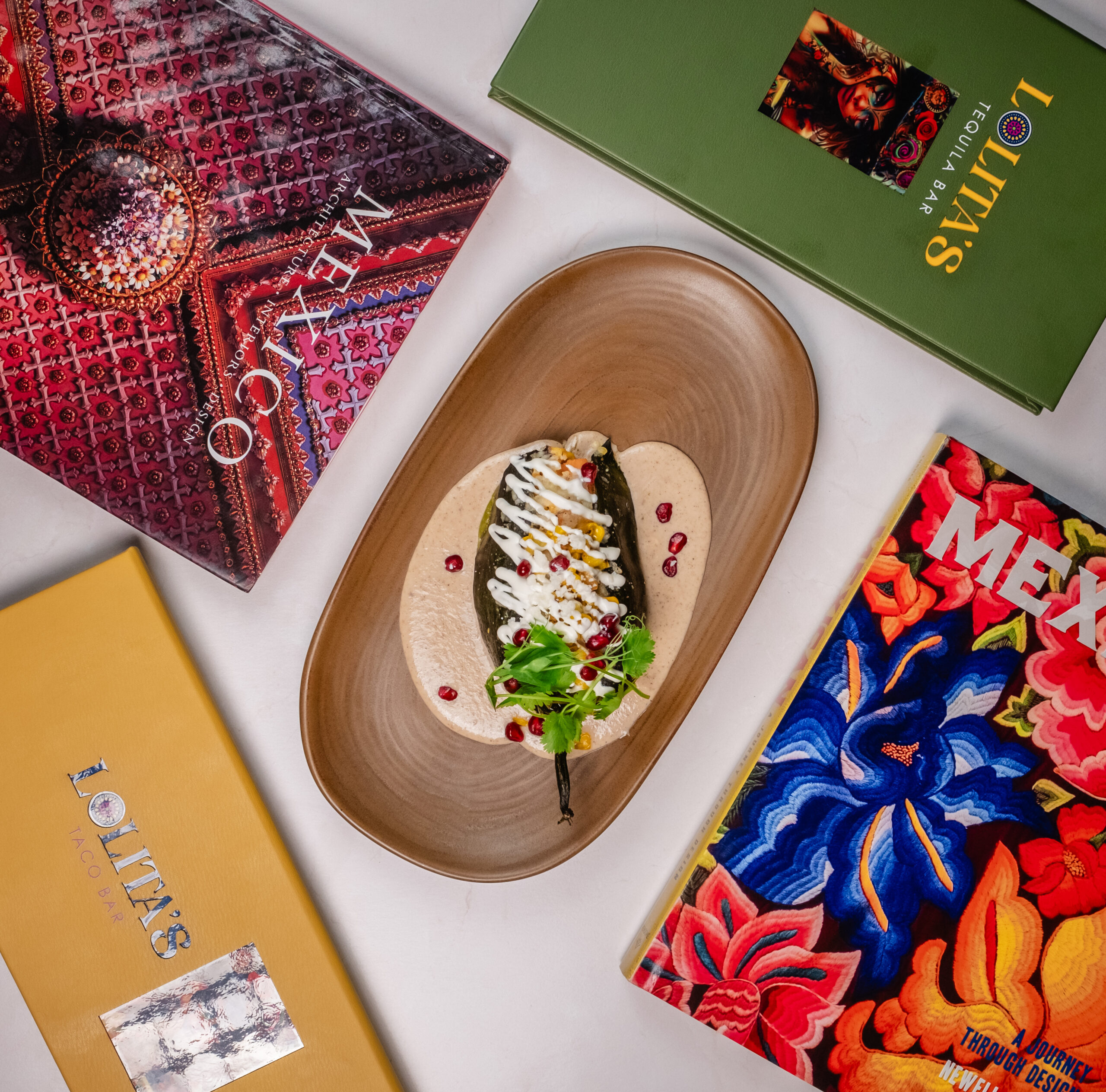 A stuffed pepper on a tortilla is garnished with sauce, herbs, and pomegranate seeds. The plate is surrounded by colorful books with covers featuring Mexican art and design.