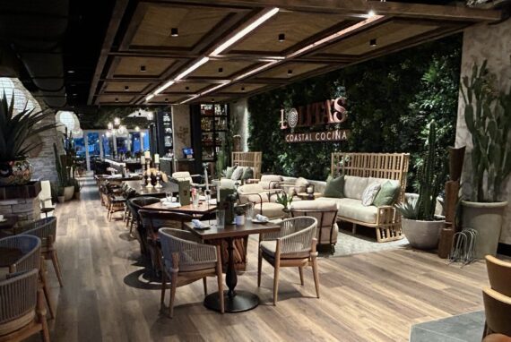 A stylish restaurant interior with wooden floors, lush green plants on the wall, cozy seating, and tables set for dining, creating a warm and inviting atmosphere.