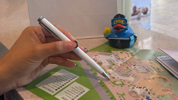 A hand holding a pen points to a colorful resort map, with a blue rubber duck wearing diving gear placed nearby on the counter.
