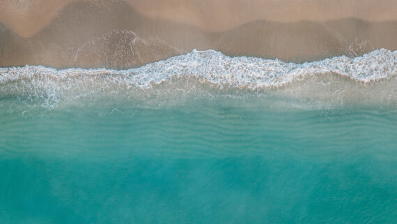 Aerial view of turquoise ocean waves gently washing onto a sandy beach, with soft white foam at the shoreline.
