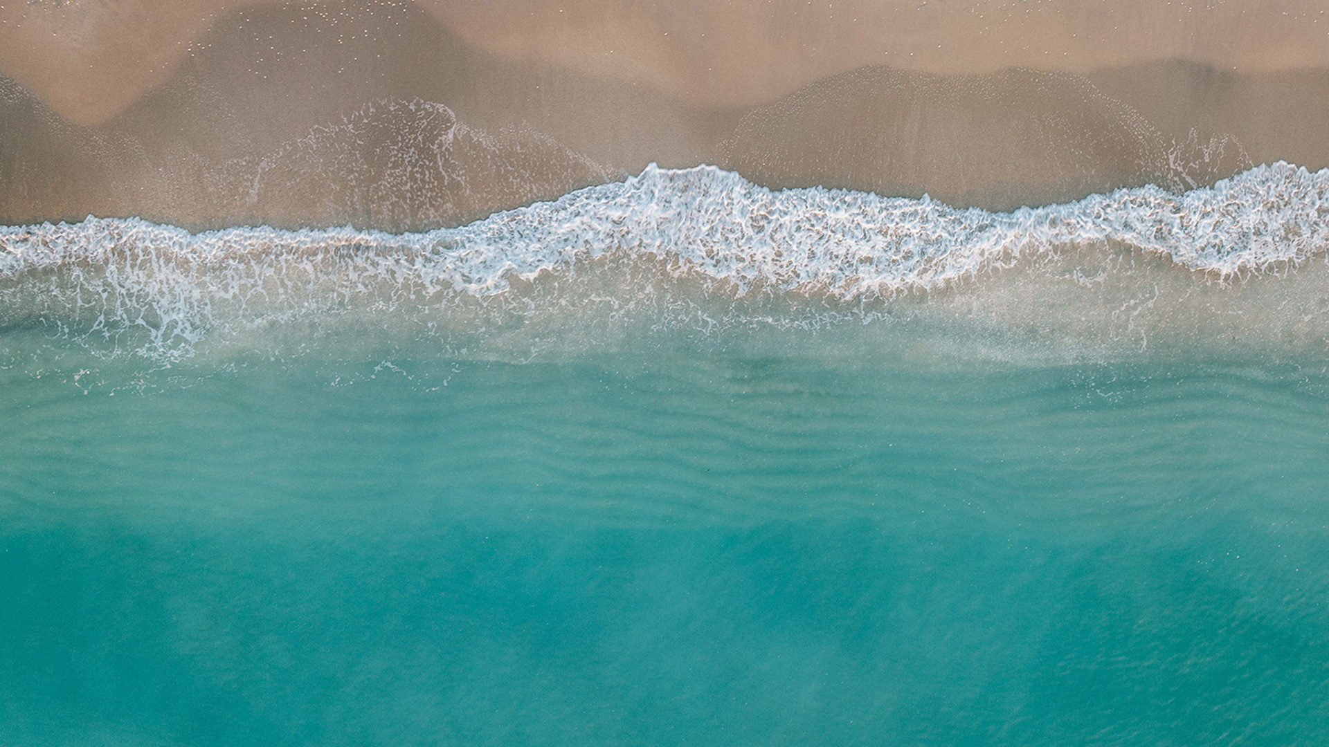 Aerial view of turquoise ocean waves gently washing onto a sandy beach, with soft white foam at the shoreline.