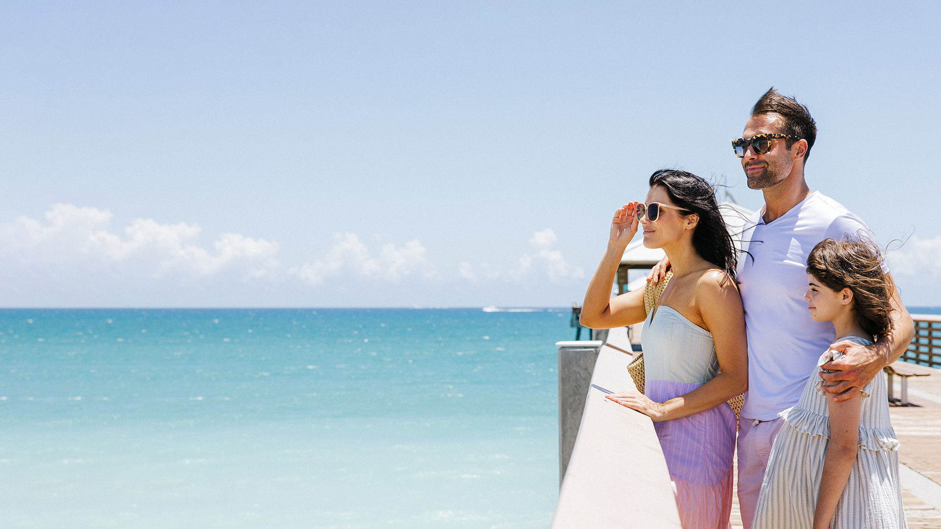 A family of three stands on a boardwalk by the ocean, looking out at the water on a sunny day. They wear sunglasses and summer clothes, with clear blue sky and turquoise sea in the background.