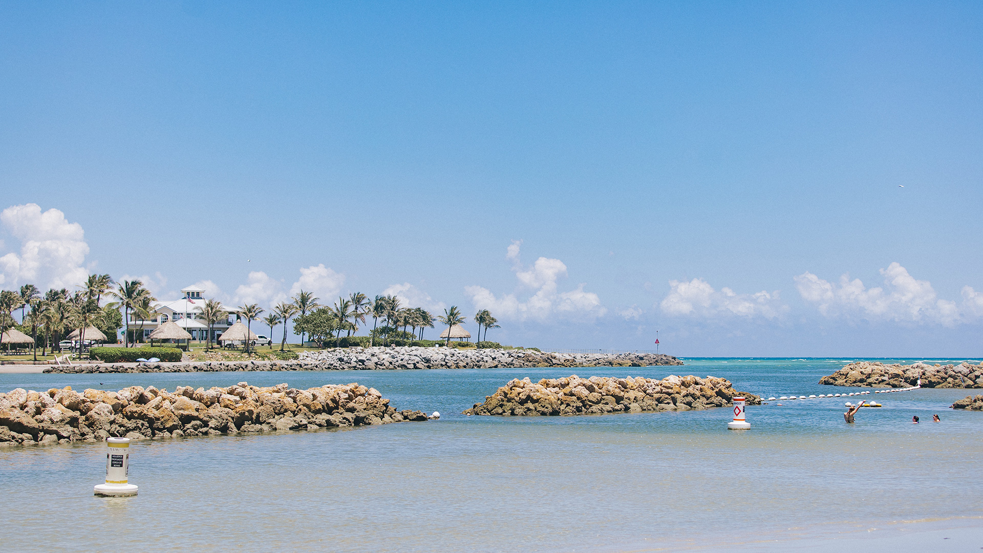 A tropical beach with clear blue water, palm trees, and rocks, under a bright sky. People are swimming and relaxing in the lagoon, while buildings and huts are visible in the background.