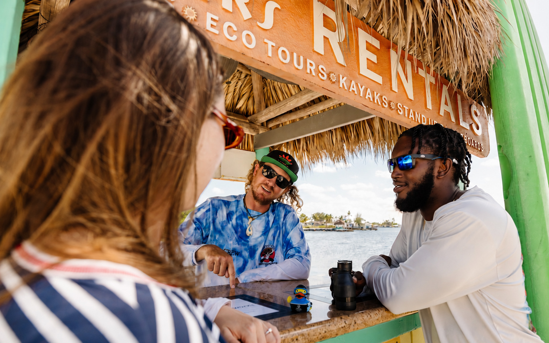 Three people talk at a shaded rental booth near the water. Two men wearing sunglasses stand behind the counter, while a woman in stripes faces them. The booth advertises eco tours, kayaks, and rentals.