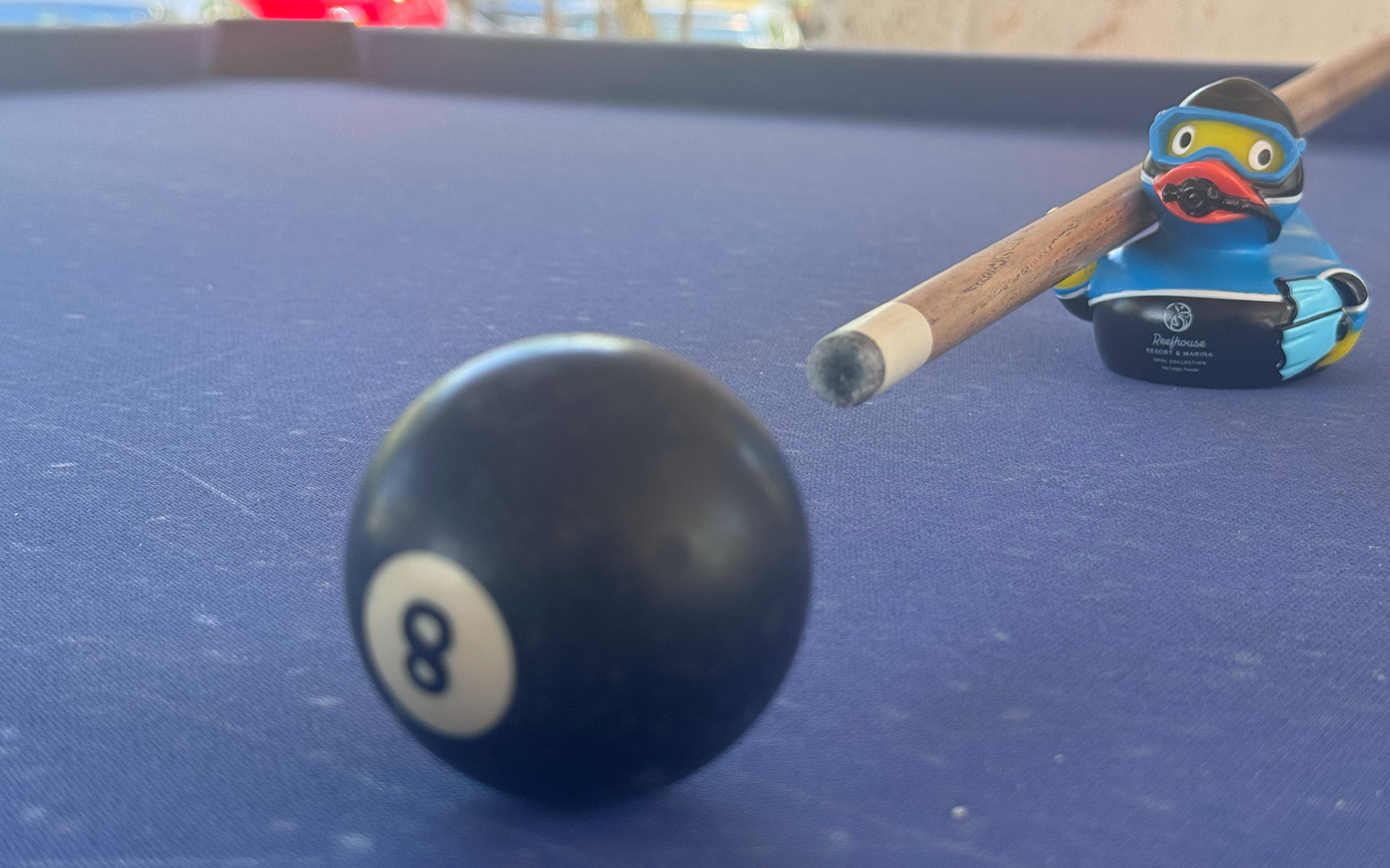 A black 8-ball sits on a blue pool table, with a pool cue and a rubber duck wearing goggles and a helmet in the background.