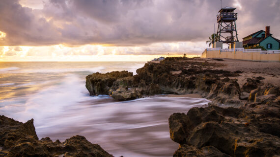 Rocky shoreline at sunrise with waves washing over rocks, a lifeguard tower, house, and cloudy sky in the background.