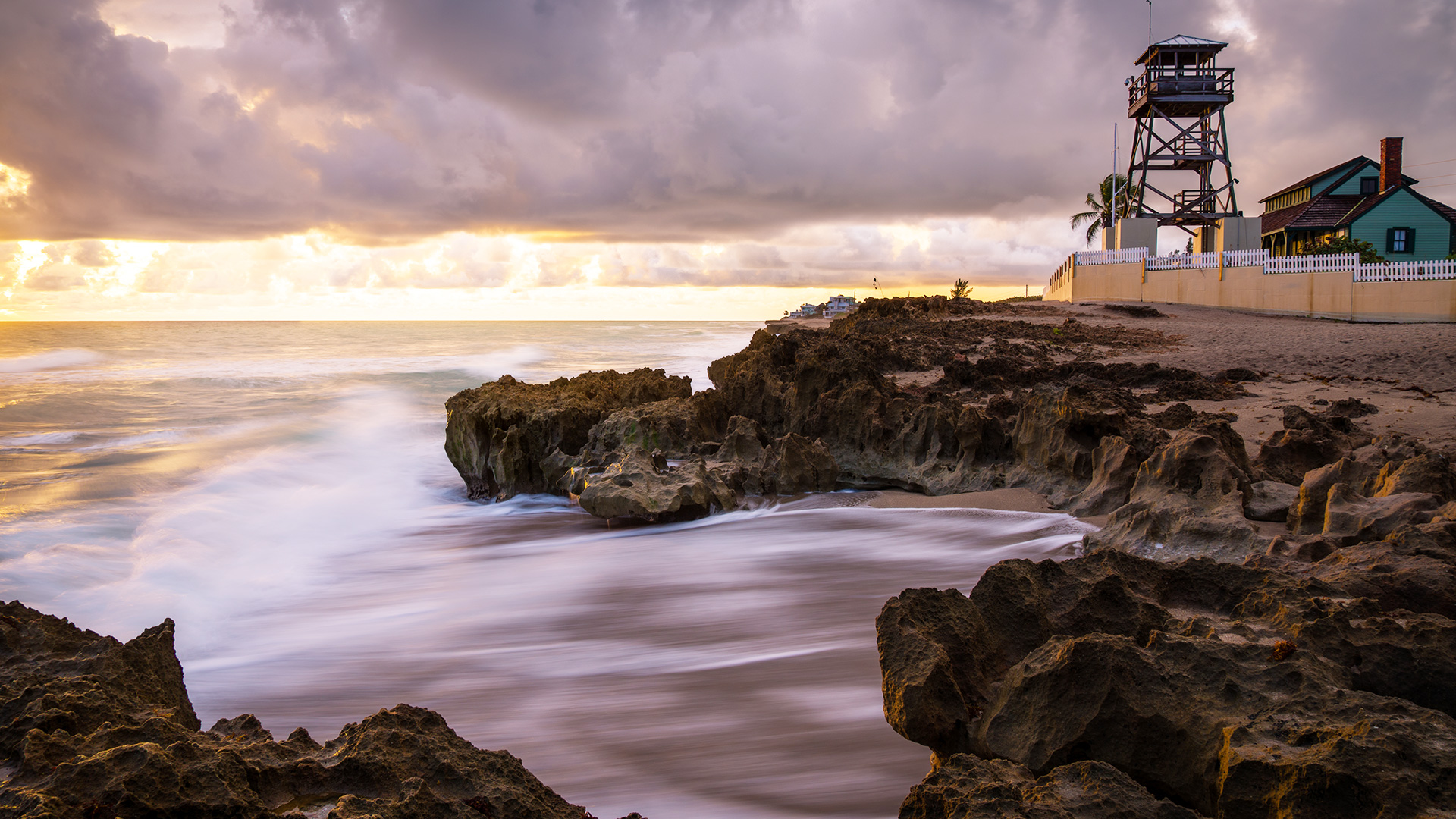 Rocky shoreline at sunrise with waves washing over rocks, a lifeguard tower, house, and cloudy sky in the background.