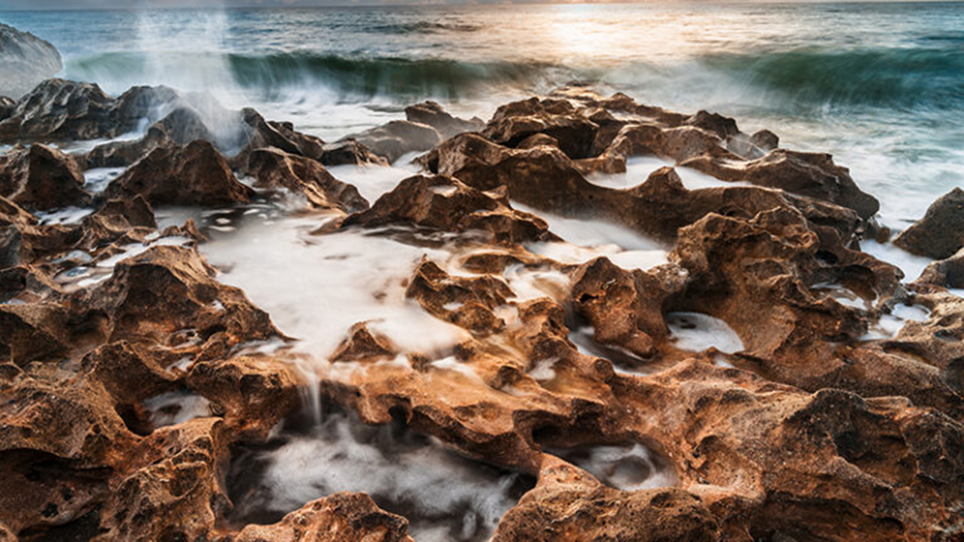 Rocky shoreline with rough, jagged rocks in the foreground and waves crashing in the background at sunset, mist rising where water meets rock.