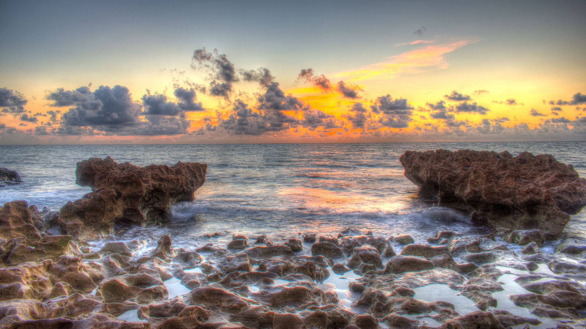 Rocky shoreline at sunrise, with jagged rocks jutting into calm ocean waters under a colorful sky filled with clouds.
