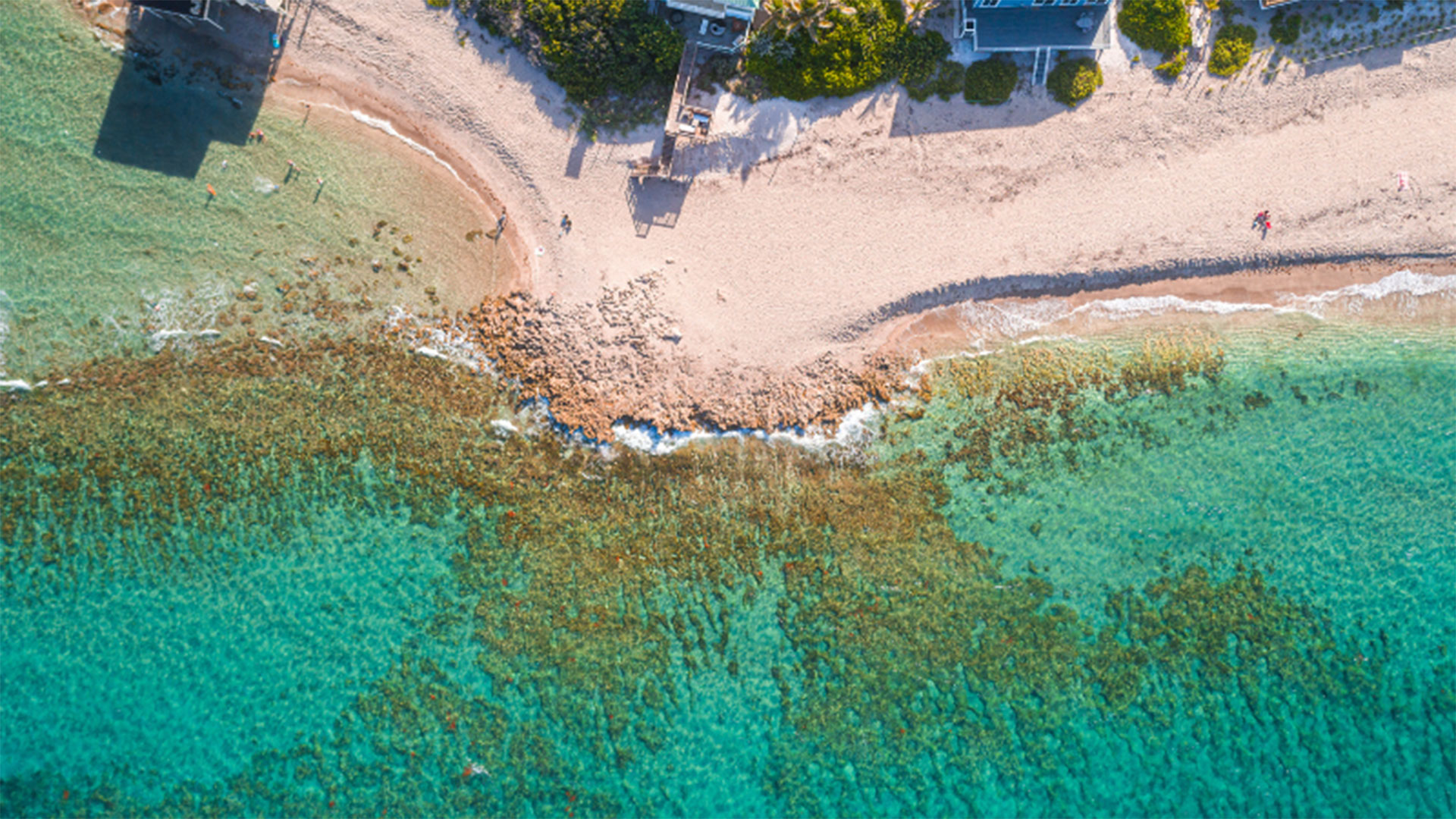 Aerial view of a sandy beach with turquoise water, a rocky shoreline, green vegetation, and a few buildings near the top. Two small figures walk along the sand.