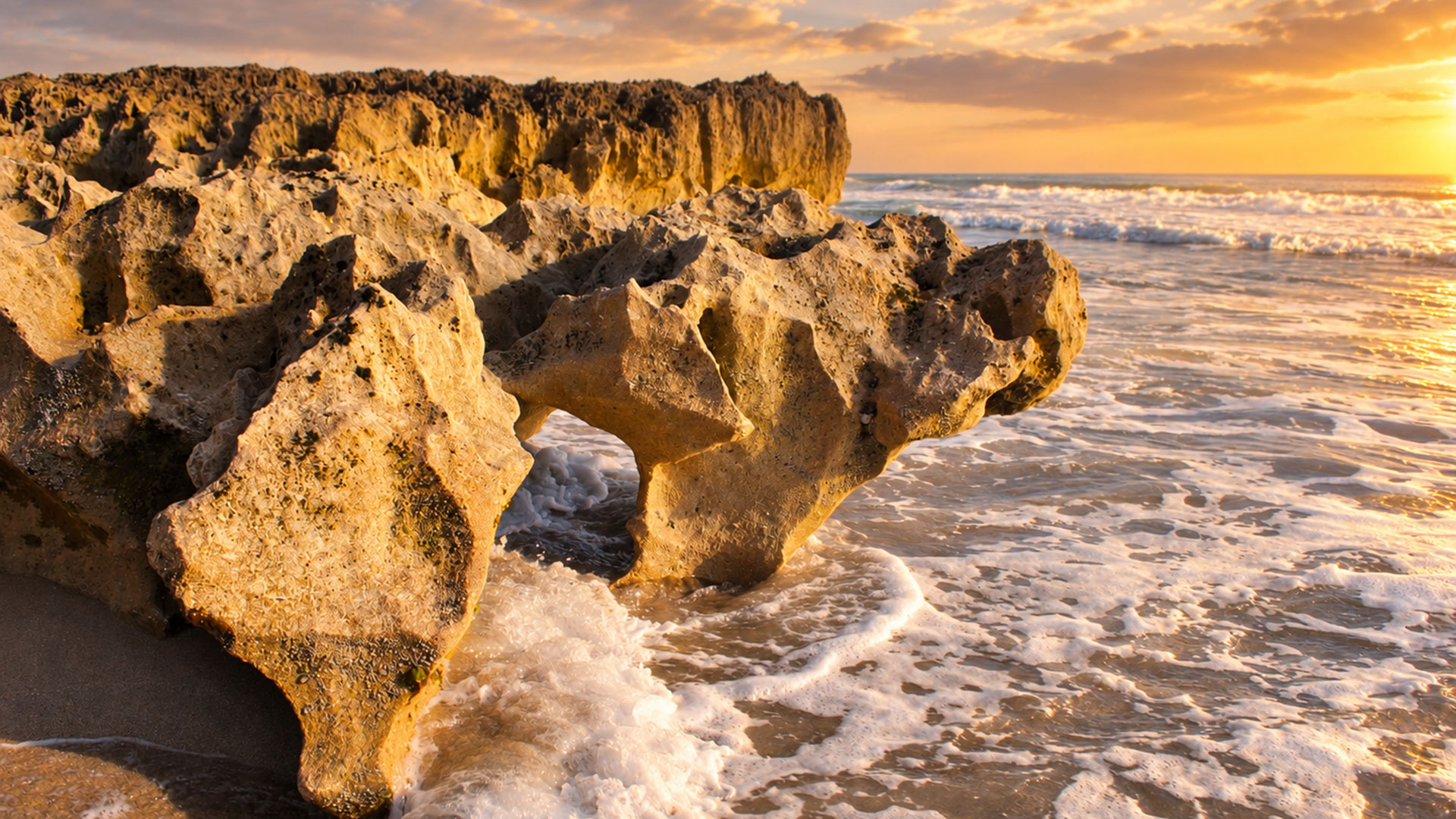 Jagged rock formations on a sandy beach with waves washing over them at sunset, golden sunlight reflecting on the water and sky.