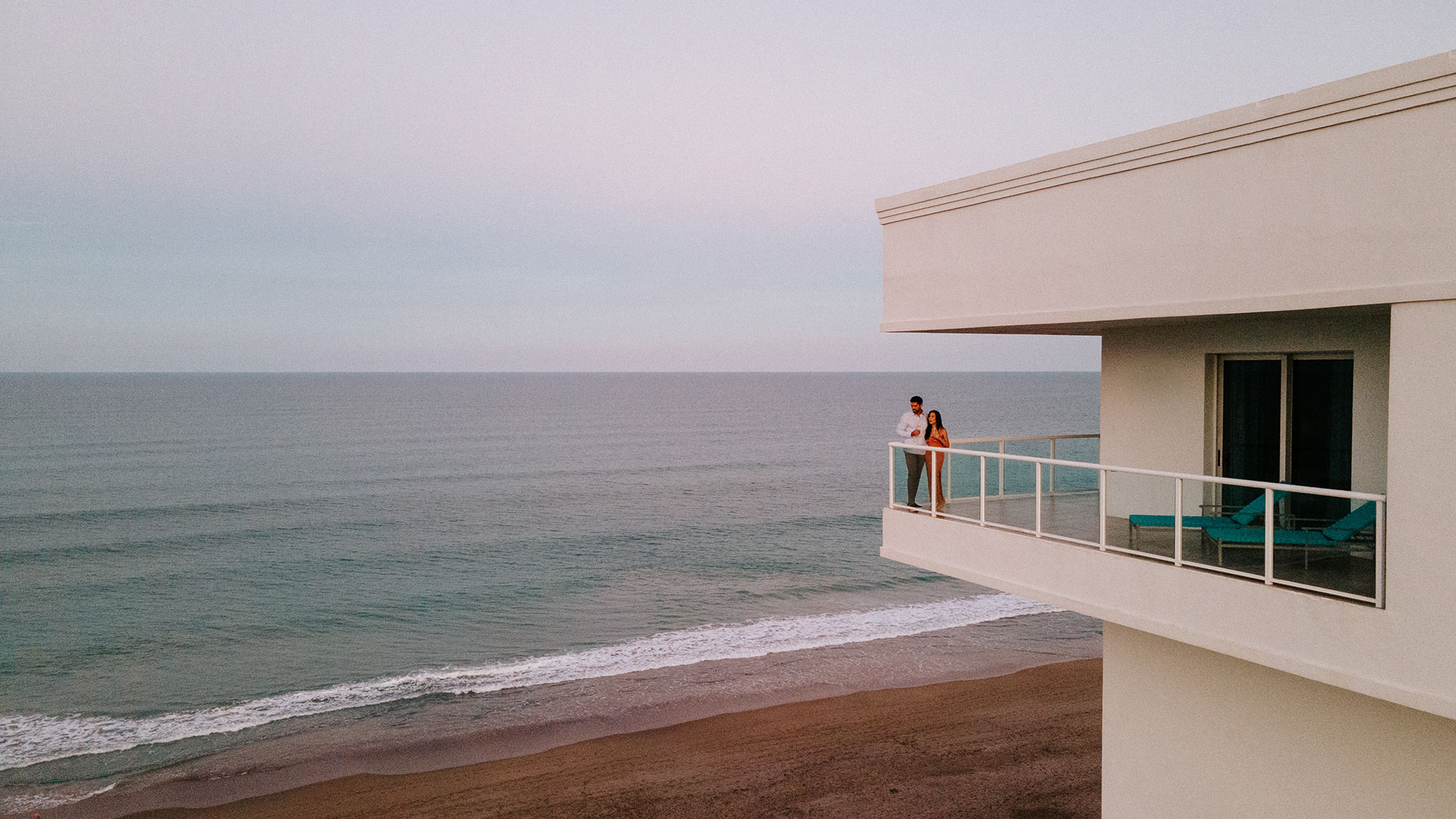 A couple stands together on a modern balcony overlooking a calm ocean and sandy beach at sunset, with soft pastel colors in the sky.