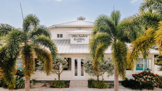 A white building labeled Olde Naples Building 1921 stands behind palm trees and bushes on a sunny day.
