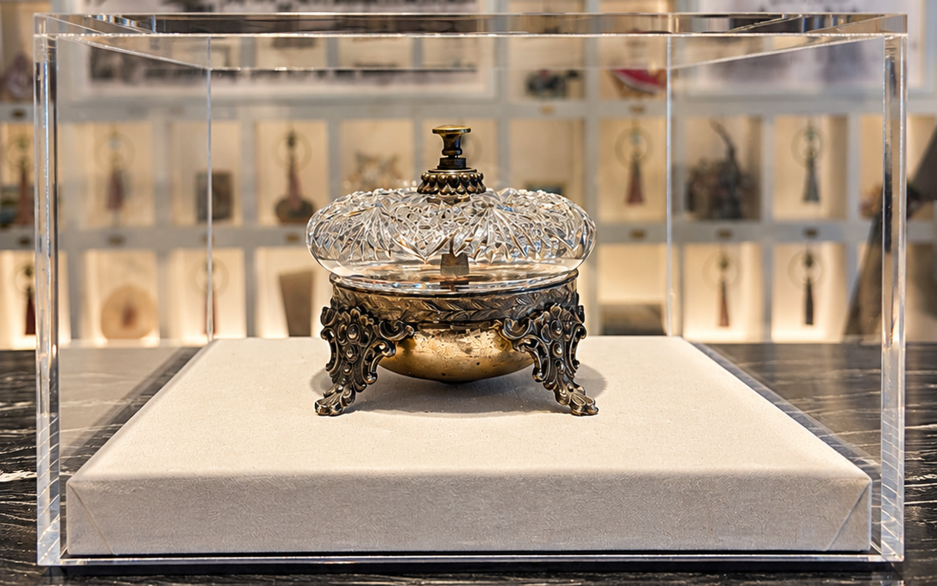 An ornate, vintage glass and metal perfume bottle is displayed on a fabric-covered pedestal inside a clear acrylic case, with a blurred background of shelves featuring various decorative items.