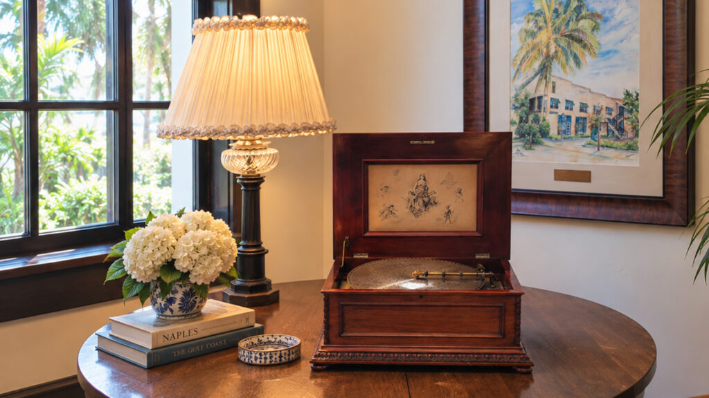 A vintage music box sits on a round wooden table beside a lamp, a vase of white flowers, stacked books, and a framed painting in a cozy sunlit room.