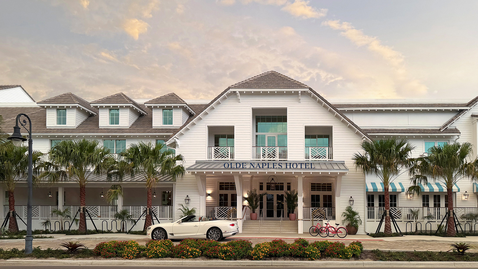 A white, coastal-style hotel with palm trees, a parked white convertible, and two red bicycles by the entrance. The building has large windows, balconies, and a sign reading Olde Naples Hotel..