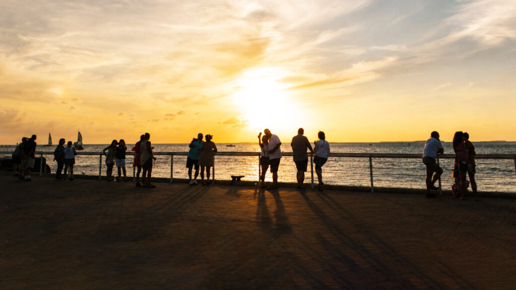 People stand along a seaside boardwalk, watching a bright yellow sunset over the ocean, with long shadows cast on the ground and sailboats visible in the distance.
