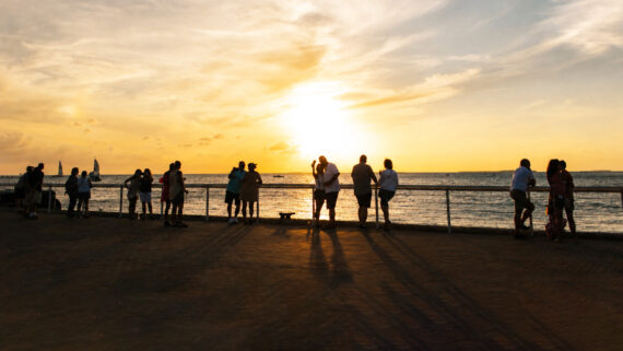 People stand along a seaside boardwalk, watching a bright yellow sunset over the ocean, with long shadows cast on the ground and sailboats visible in the distance.