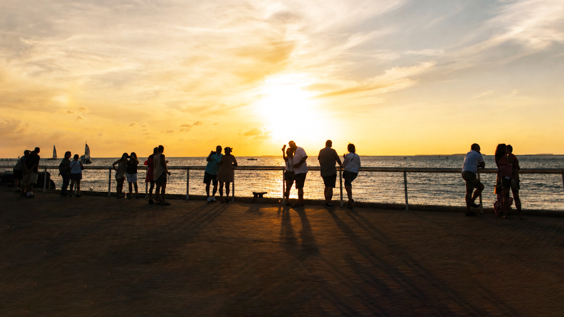 People stand along a seaside boardwalk, watching a bright yellow sunset over the ocean, with long shadows cast on the ground and sailboats visible in the distance.