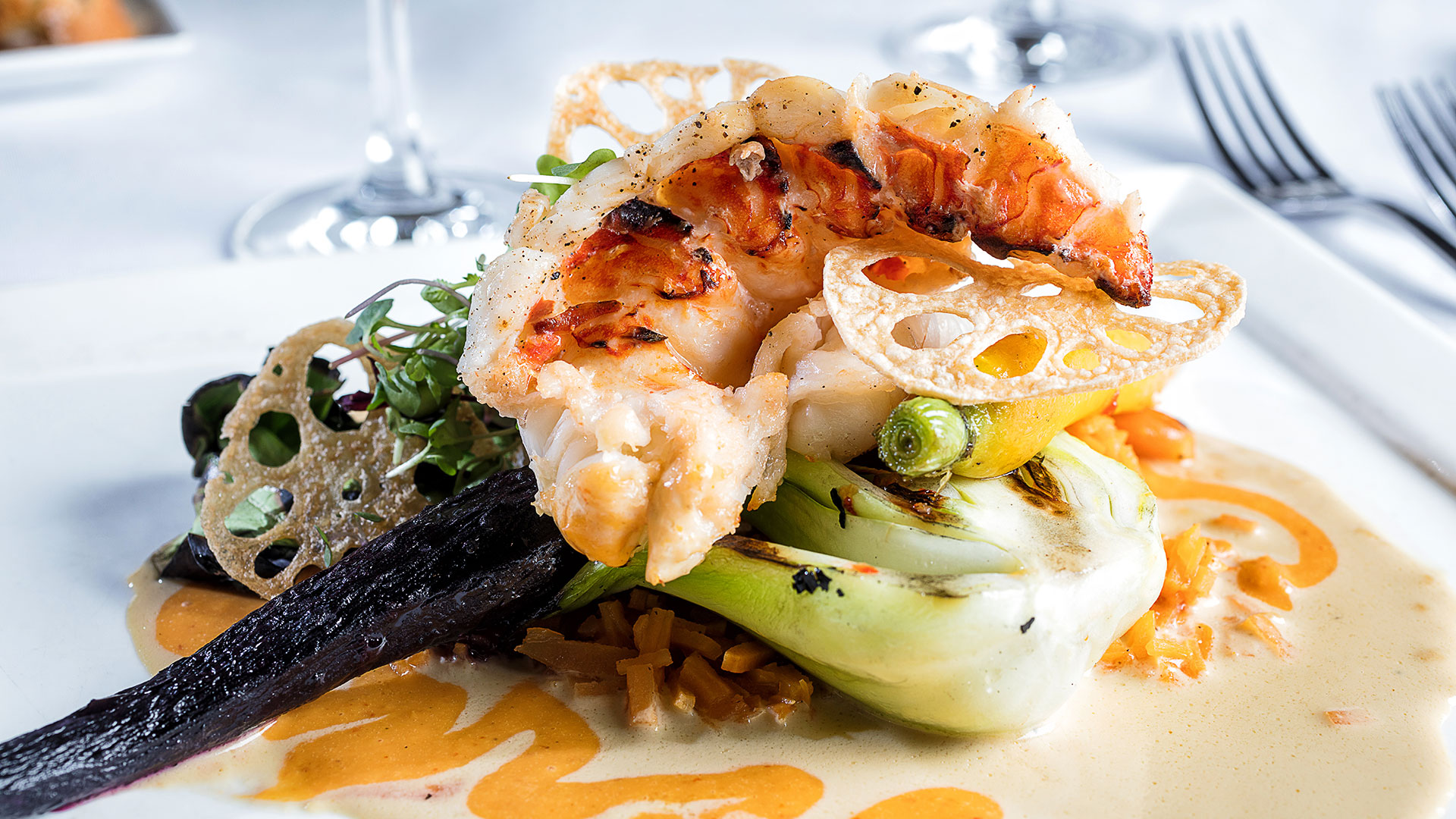 Grilled lobster tail atop bok choy and vegetables, garnished with a crispy lotus root chip, served on a white plate with a creamy orange sauce.