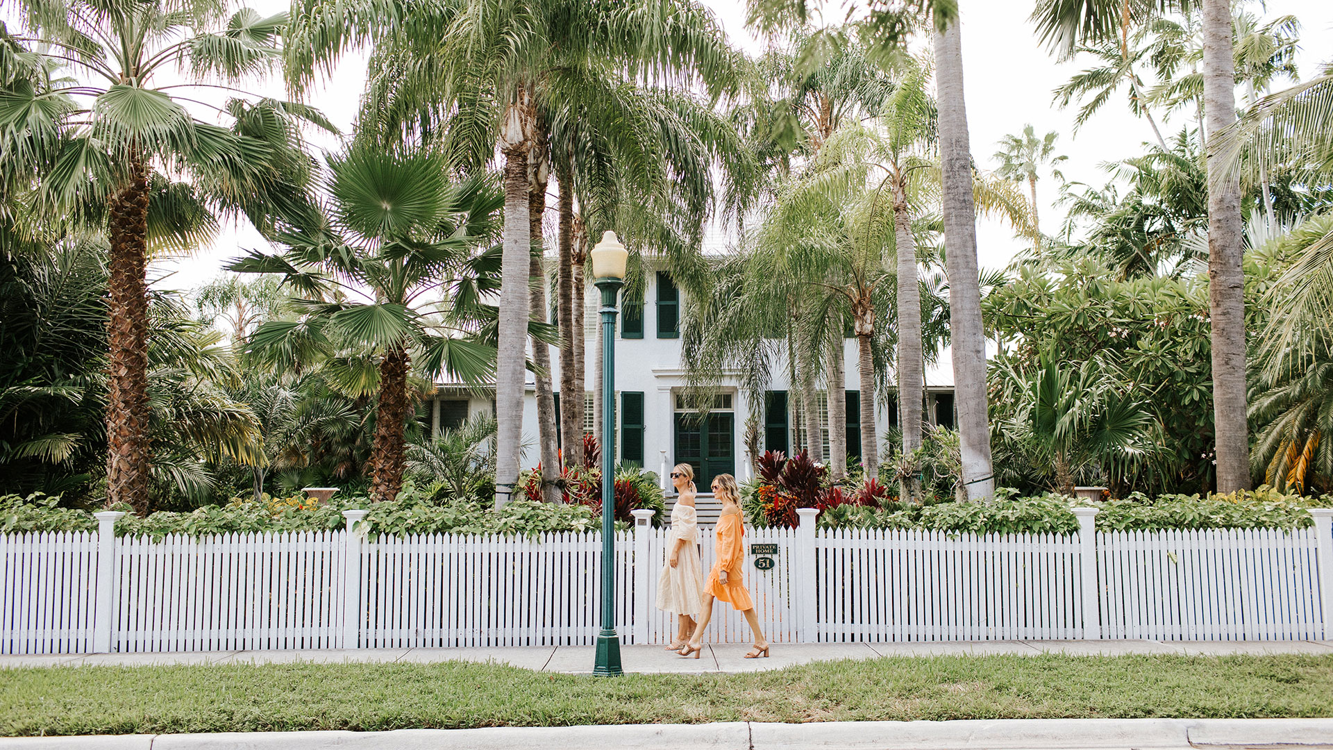 Two women walk on a sidewalk in front of a white picket fence and a house surrounded by tall palm trees and lush greenery.