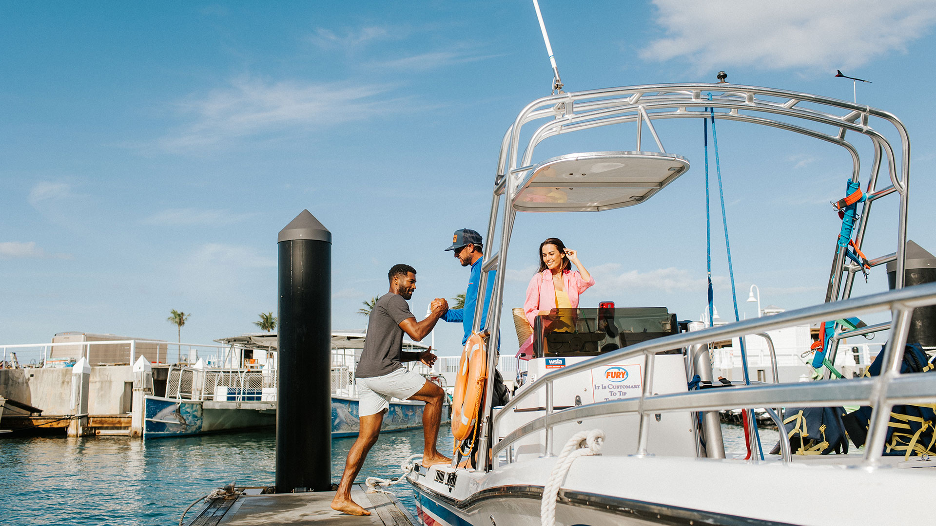 A man steps onto a boat from the dock, assisted by another man, while a woman in pink sits on the boat smiling under a sunny blue sky.