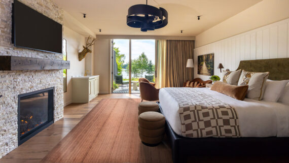 Modern hotel room with a large bed, fireplace, wall-mounted TV, armchairs, and glass doors leading to a balcony with trees and a lake view. Decor includes a geometric rug and elegant lighting.
