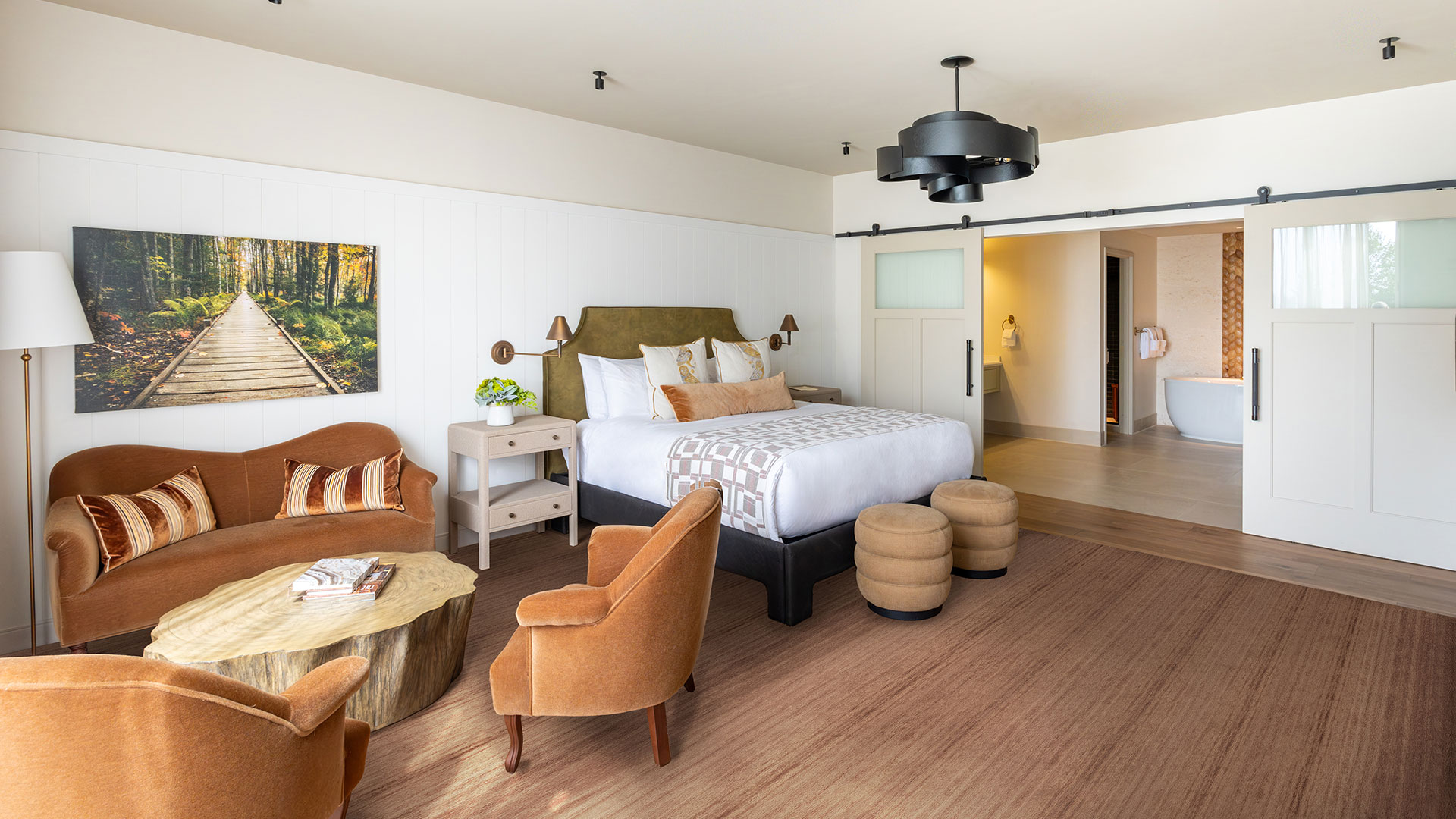 A modern hotel room with a bed, two brown chairs, a loveseat, a round wooden coffee table, and a large photo of a forest on the wall. Sliding doors reveal a bathroom with a bathtub.